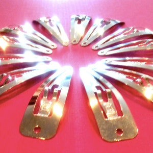 Puede incluir: Un conjunto de 12 pinzas para el pelo plateadas dispuestas en círculo sobre un fondo rosa. Las pinzas son todas del mismo tamaño y forma, y tienen un acabado brillante.