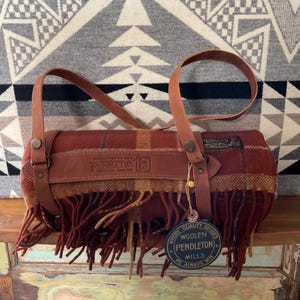 May include: A rolled-up Pendleton wool blanket with leather straps and handles. The blanket features a plaid pattern in shades of red, brown, and yellow, with a fringed edge. The leather straps are a light brown color. A tag reads "Pendleton Woolen Mills."