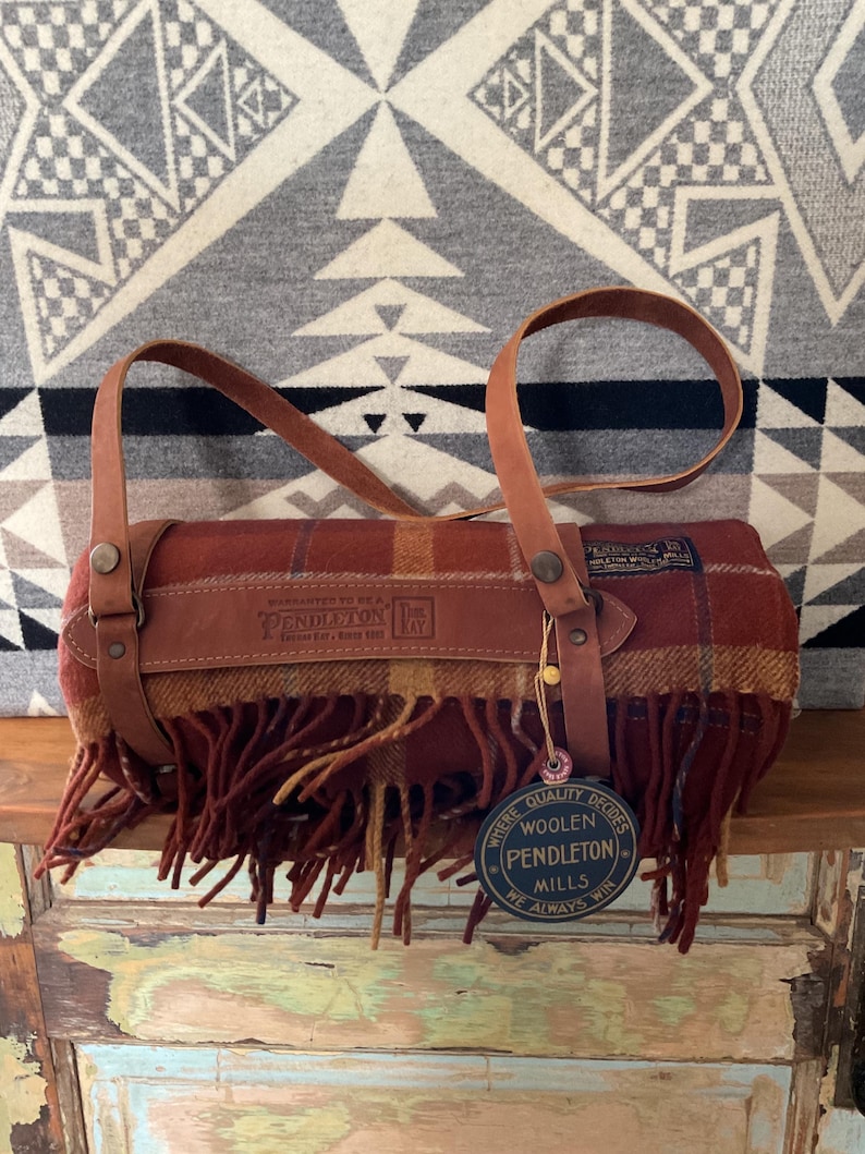 May include: A rolled-up Pendleton wool blanket with leather straps and handles. The blanket features a plaid pattern in shades of red, brown, and yellow, with a fringed edge. The leather straps are a light brown color. A tag reads "Pendleton Woolen Mills."