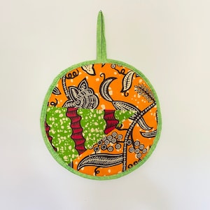 May include: Round orange and green fabric potholder with a loop for hanging. The potholder features a colorful floral and leaf pattern with a green border and a green loop. The design includes black, white, and red accents.