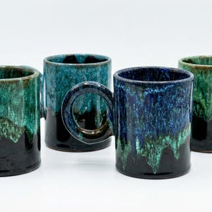 May include: Four ceramic mugs with a unique glaze. The mugs have a dark base that transitions to shades of green and blue. Each mug has a large, rounded handle. The mugs are displayed on a white background.