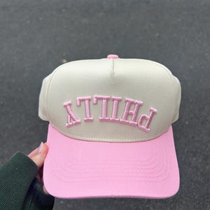 May include: A baseball cap with a light beige crown and a pink brim. The word "PHILLY" is embroidered in pink across the front of the cap. The cap is made of a soft fabric.