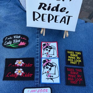 May include: A collection of embroidered patches featuring various designs and text including Lady Biker, Lady Rider, Hope with a pin-up illustration, Does This Bike Make My Ass Look Fast, and I Just Look Sweet & Innocent, displayed with a sign reading SLEEP, RIDE, REPEAT.