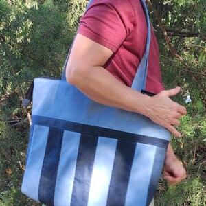 May include: A blue tote bag with vertical stripes in varying shades of blue and black. The bag has a rectangular shape with a zippered top and long shoulder straps. The bag is being carried over the shoulder.