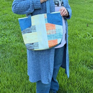 May include: A tote bag featuring a patchwork design with panels of blue denim, light yellow fabric, and a textured orange and blue patterned section. The bag has two matching blue denim shoulder straps and a rectangular shape with a flat base.