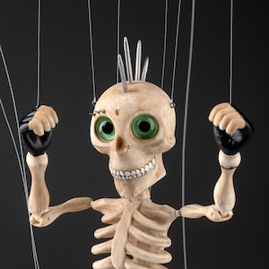 May include: A skeleton marionette puppet with a beige bone-colored finish, large green eyes, and a silver mohawk. The figure wears black gloves and black boots, with small black cross symbols on the knees and white strings attached for movement.