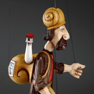 May include: A wooden puppet of a person wearing a brown jacket, red shirt, and striped socks. The puppet has a snail shell on its head and a snail shell backpack with a small white house on top. The puppet is walking with its arms outstretched.