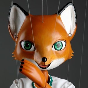 May include: A fox puppet with a white and orange face, green eyes, and a white shirt with a green trim. The puppet is holding its arms in front of its chest and is wearing a green skirt.