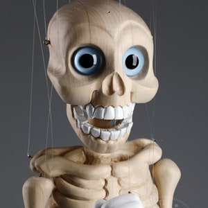 May include: A wooden skeleton puppet with a detailed, carved skull featuring blue eyes and a wide, toothy grin. The puppet's arms are posed, and it has a white hand. Strings are attached for manipulation, suggesting a toy or decorative item.