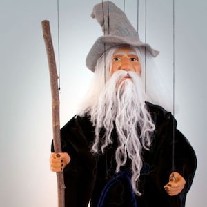 May include: A handcrafted wizard marionette with a long white beard, gray hat, and black robe. The puppet holds a wooden staff and is suspended by strings. The marionette is a Czech-made original.