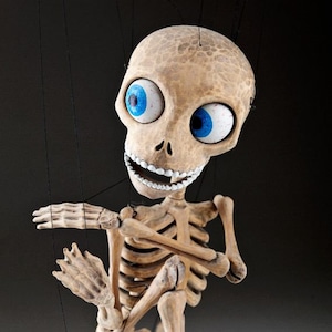 May include: A wooden skeleton puppet with large blue eyes, a toothy grin, and black and white shoes. The puppet is posed in a dancing position with its arms crossed and one leg bent. The puppet is attached to strings.
