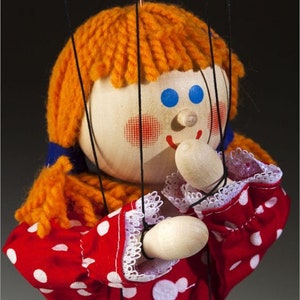 May include: A red and white polka dot puppet with a white apron and blue skirt. The puppet has orange yarn hair, blue eyes, and a painted smile. The puppet is made of wood and has strings attached to its arms and head.