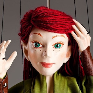 May include: A red-haired puppet with green eyes and freckles, wearing a green shirt and brown leather belt. The puppet is holding its hand up to its head, as if adjusting its hair.