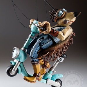 Puede incluir: Un títere de madera de un perro con gafas y un chaleco vaquero, montando un scooter turquesa. El títere está hecho a mano y tiene una estética rústica y vintage.