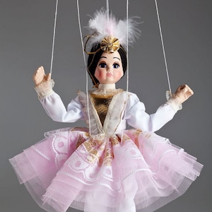 May include: A pink and white ballerina puppet with a gold bodice and a white feather headdress. The puppet is wearing white ballet shoes and is posed in a dancing position. The puppet is made by Czech Marionettes and is hand-made.