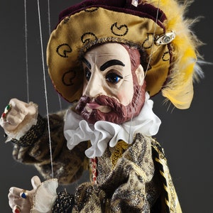 Op de afbeelding: Een pop met een bruine baard en snor, die een gouden en bordeauxrode hoed draagt met een gele veer, een witte geplooide kraag en een bruin en gouden gepatroneerd jasje.