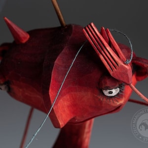 Puede incluir: Un títere de madera rojo con una nariz larga y puntiaguda y ojos grandes y expresivos. El títere lleva un sombrero rojo con una punta puntiaguda y está sujeto a una cuerda.