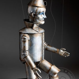 May include: A silver-colored, tin man puppet with a painted face and a conical hat. The puppet is kneeling with its right arm extended and its left hand resting on its knee. The puppet is made of metal and has a detailed, realistic appearance. The puppet is attached to strings, suggesting it is a marionette.