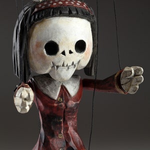 May include: A wooden puppet with a skull-like face, red and white polka dot headband, and red and black clothing. The puppet has black hair and is holding its arms out.