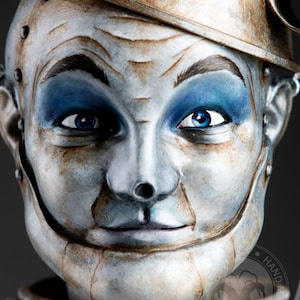 May include: Close-up of a painted metal sculpture of a human face with blue eyes and a painted metal hat. The sculpture has a distressed, aged look with visible brushstrokes.