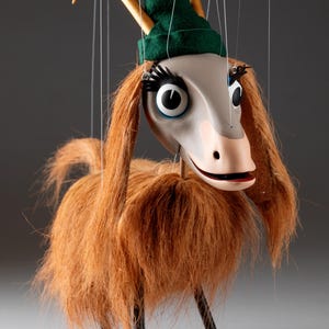 Puede incluir: Un títere artesanal de una cabra con pelo largo marrón, que lleva un sombrero verde con un cuerno dorado. El títere tiene grandes ojos negros y una nariz rosa.