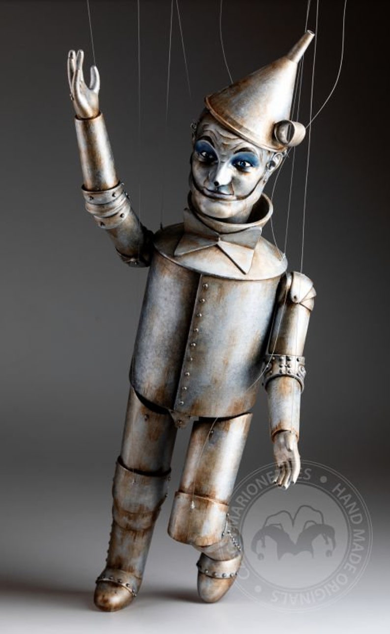 May include: A hand-crafted, vintage-style tin man puppet with a funnel hat, painted details, and a distressed metal finish. The puppet is posed with one arm raised and the other extended, with strings attached to its limbs. The puppet is standing on a gray surface. The image includes the text "MARIONETTES HAND MADE ORIGINALS."