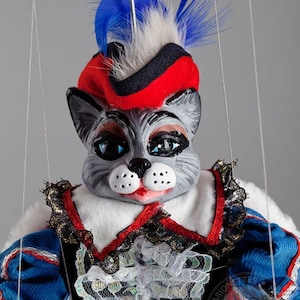 May include: A gray cat puppet wearing a red and black hat with a blue feather. The puppet is dressed in a blue and white costume with a white lace collar. The puppet is being controlled by strings.