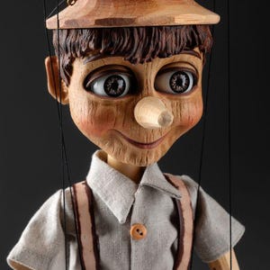 May include: A wooden marionette puppet featuring a carved face with a long nose, large brown eyes, and a light brown hat. The puppet wears a light gray short-sleeved shirt with two buttons and brown suspenders, suspended by thin black control strings.
