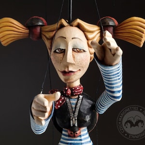 May include: A wooden puppet of a girl with blonde pigtails, wearing a blue and white striped shirt, a red and white polka dot scarf, and a black waistcoat with a zipper. The puppet is pointing with both hands and has a friendly expression.