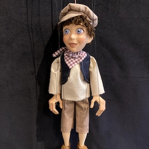 Puede incluir: Un títere de madera hecho a mano de un niño que lleva una gorra marrón, una camisa blanca, un chaleco azul, pantalones marrones y zapatos marrones. El títere tiene el pelo castaño, ojos azules y una bufanda a cuadros rojos y blancos.