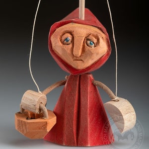 May include: A wooden puppet of Little Red Riding Hood, with a red hooded dress, a basket, and a sad expression. The puppet is made by Czech Marionet and is hand-made.