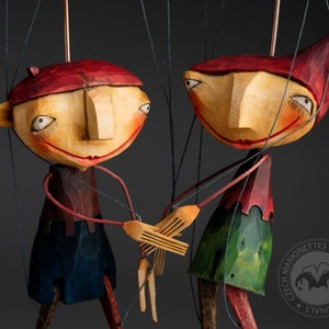 Puede incluir: Dos marionetas de madera con caras pintadas y ropa colorida. Las marionetas se toman de la mano y están suspendidas por cuerdas. Las marionetas están hechas por Czech Marionettes Originals y son hechas a mano.