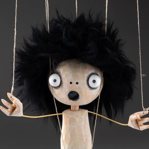 Puede incluir: Una marioneta con cabeza y torso de madera tallada en color claro con una superficie texturizada. La marioneta tiene grandes ojos redondos blancos con pupilas oscuras, una boca pequeña y oscura, y una espesa y voluminosa cabellera negra.