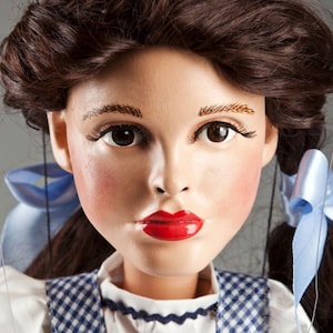 Puede incluir: Una muñeca de porcelana con cabello castaño peinado en dos trenzas con cintas azules. La muñeca lleva una blusa blanca y un vestido de cuadros azules y blancos.