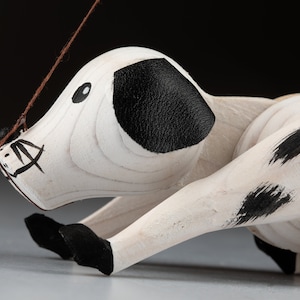 May include: A wooden toy dog with a white body and black spots, ears, and feet. The dog has a painted face with a black eye and a brown string attached to its head. The toy is lying down.