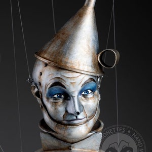 May include: A hand-crafted puppet of the Tin Man from the Wizard of Oz. The puppet is made of painted wood and metal, and has a funnel-shaped hat. The puppet's face is painted with a realistic expression, and it has blue eyes.