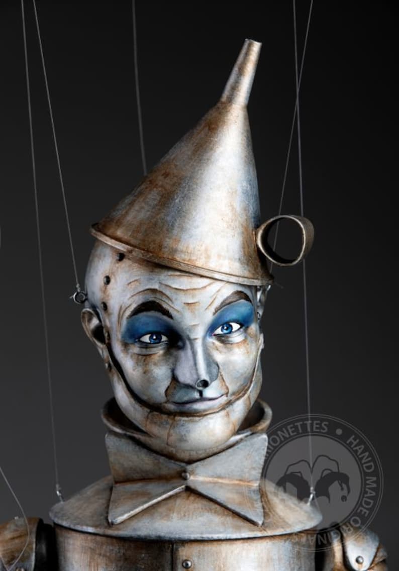 May include: A hand-crafted puppet of the Tin Man from the Wizard of Oz. The puppet is made of painted wood and metal, and has a funnel-shaped hat. The puppet's face is painted with a realistic expression, and it has blue eyes.