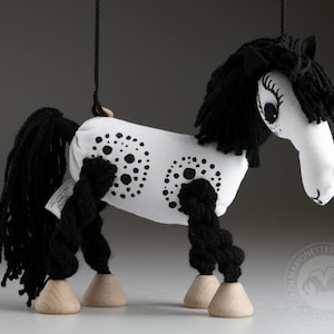 Puede incluir: Un caballo de títeres blanco y negro con una larga melena y cola negras. El caballo tiene lunares negros en su cuerpo blanco y pies de madera.