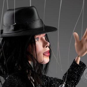 May include: A black and white puppet with a black fedora hat, black jacket, and long black hair. The puppet is wearing a black and white patterned jacket. The puppet is holding its right hand up in the air. The puppet is being controlled by strings.
