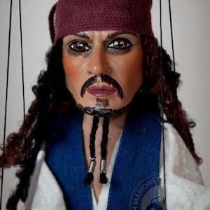Puede incluir: Un títere artesanal de Captain Jack Sparrow de las películas de Piratas del Caribe. El títere está vestido con una camisa azul y blanca, pantalones marrones y una bandana roja. El títere tiene el pelo largo y negro y barba, y sostiene una espada.