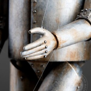 May include: Close-up of a hand made of metal and wood, with a string attached to it. The hand is part of a larger figure, possibly a robot or a puppet. The figure is made of metal and has a weathered, rusty appearance.
