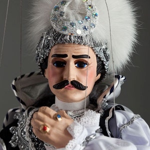 Puede incluir: Títere detallado de un hombre con un gran bigote, vestido con un atuendo blanco y plateado con un tocado de plumas y una media luna enjoyada. El títere tiene ojos azules y junta las manos, adornadas con anillos.