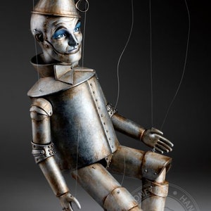 May include: A silver and rust-colored tin man puppet with blue eyes and a painted smile. The puppet is suspended by strings and has a conical hat. The puppet is sitting with its legs crossed and its arms outstretched. The text "HAND MADE" is visible on the bottom right of the image.