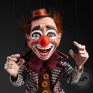Puede incluir: Un títere artesanal de payaso con nariz roja, maquillaje blanco y chaqueta a cuadros. El títere lleva un sombrero rojo y negro y tiene una gran sonrisa.