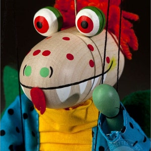 May include: A wooden puppet with a red, green, and white face, wearing a blue and black polka dot shirt and a yellow shirt underneath. The puppet has a large, toothy grin and a red tongue sticking out. The puppet is holding a green hand up in the air. The puppet is made by Czech Marionettes Hand Made Originals.