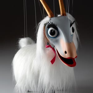 Puede incluir: Una marioneta de cabra caprichosa con una cabeza gris, ojos grandes y largas pestañas. Tiene cuernos dorados, una nariz rosa y un lazo rojo. El cuerpo de la marioneta está cubierto de pelaje blanco y tiene patas negras.
