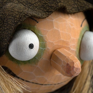 May include: Close-up of a hand-carved wooden puppet head. The puppet has large white eyes with black pupils, a long nose, and a textured, orange face. It wears a dark brown hood and has straw-like hair.