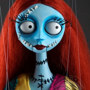 Puede incluir: Un títere azul y blanco con cabello rojo, costuras cosidas y ojos grandes. El títere lleva un vestido de retazos colorido con un patrón rosa y negro. El títere está suspendido por cuerdas.