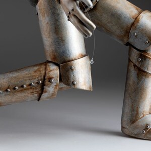 May include: A silver and brown metal puppet of a robot with articulated legs and arms. The robot is in a running pose with one leg forward and the other leg back. The robot is made of metal and has a weathered finish.