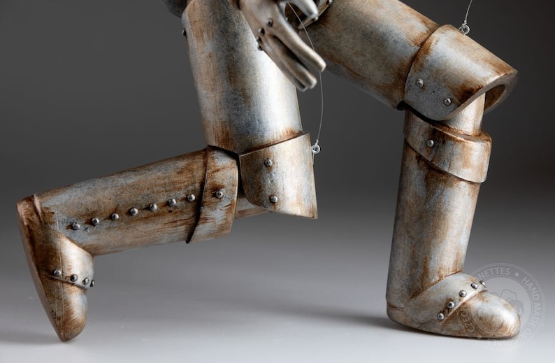 May include: A silver and brown metal puppet of a robot with articulated legs and arms. The robot is in a running pose with one leg forward and the other leg back. The robot is made of metal and has a weathered finish.